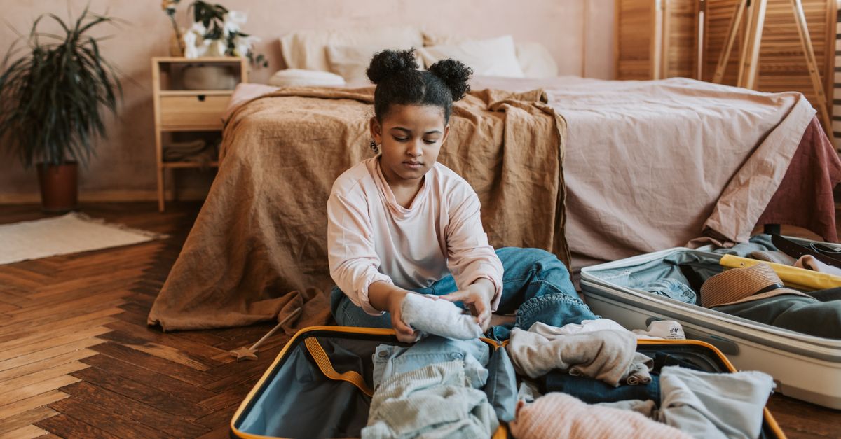 A young girl folding clothes into a suitcase in a cozy bedroom setting with natural decor.